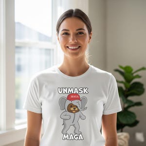 May include: White t-shirt with the words "UNMASK MAGA" in black block letters. The shirt features a cartoon elephant wearing a red hat with "MAGA" on it. The elephant is dressed in a gray elephant costume.