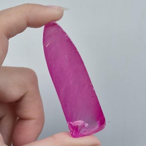 May include: A vibrant, elongated, pink gemstone with a smooth, polished surface. The gemstone is held between fingers, showcasing its translucent quality and irregular, fractured edge. The colour is a deep, saturated pink.