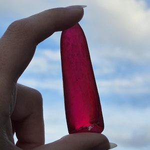 May include: A translucent, elongated, deep magenta-colored gemstone is held between fingers against a backdrop of a cloudy blue sky. The gem has a smooth, polished appearance with a slightly irregular shape.
