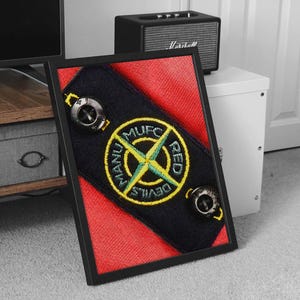 May include: A framed print of a black and red patch with a yellow circle and the text "RED DEVILS MANU MUFC" in a green and yellow font.