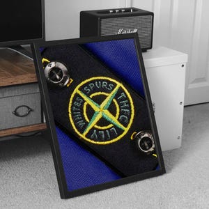 Puede incluir: Una impresión enmarcada de una tela negra y azul con un parche bordado amarillo y verde. El parche es una brújula con las palabras "Lily White Spurs THFC" alrededor.