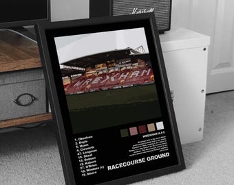Può includere: Una stampa incorniciata raffigurante lo stadio Racecourse Ground con la parola "WREXHAM". La stampa include un elenco di nomi e numeri e il testo "RACECOURSE GROUND". La cornice è nera.