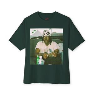 Könnte beinhalten: Ein dunkelgrünes T-Shirt mit einem aufgedruckten Bild einer Person, die ein weißes Bandana mit schwarzen Designs trägt, in einem Fahrzeug sitzt und eine Wasserflasche und eine Zigarette hält. Die Person trägt ein weißes T-Shirt und eine dunkle Hose.