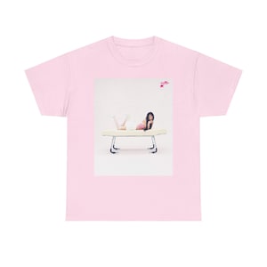 Puede incluir: Una camiseta rosa con un gráfico de una mujer tumbada en un banco blanco. La mujer lleva una falda blanca y una blusa rosa. El gráfico está en un marco blanco.