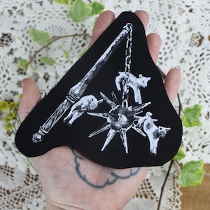 May include: Black and white embroidered patch of a spiked ball on a chain with a wooden handle and three small lambs attached to the ball.