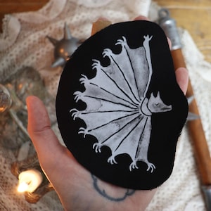 May include: A black fabric patch with a white embroidered design of a bat-like creature with a large wingspan. The creature has a long snout and a collar around its neck.