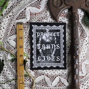 May include: A black and white patch with the text "PROTECT TRANS LIVES" in gothic lettering. The image on the patch features a person in armour. The patch is framed with a white lace border. A vintage wooden ruler and a rusty axe are also in the image.