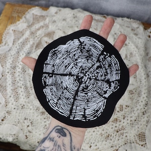 May include: Black fabric patch featuring a white tree ring design. The design resembles a cross-section of a tree trunk, showing the growth rings.