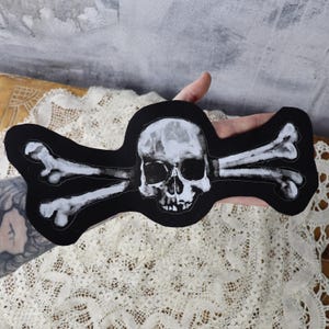 May include: A black fabric patch featuring a white skull and crossbones design. The skull has a detailed face with eye sockets, a nose, and a mouth.