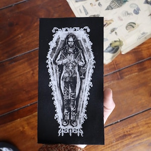 May include: Black and white embroidered patch depicting a person lying in a coffin. The person is wearing a long coat and holding a chalice. The coffin is decorated with ornate details.