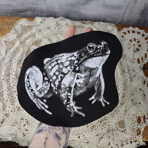 May include: Black fabric patch with a white silhouette of a frog. The frog has a spotted pattern and is facing to the right.