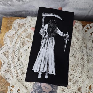 May include: A black fabric patch with a white illustration of a figure holding a scythe and a dagger. The figure is wearing a long, flowing gown and has long hair.