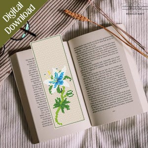 May include: Cross stitch bookmark featuring a white flower with blue petals and green leaves. The bookmark is on a book with text in Dutch.