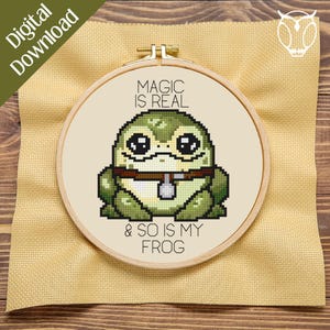 May include: A cross-stitch embroidery hoop featuring a pixelated green frog with large eyes and a brown collar. The text "MAGIC IS REAL & SO IS MY FROG" is stitched above and below the frog. The background is a light beige fabric.