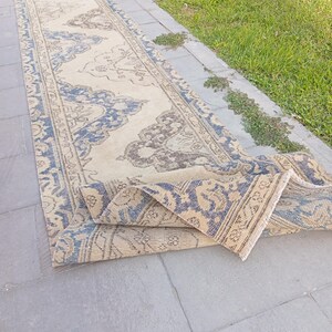 May include: A long, narrow runner rug featuring a cream-coloured base with intricate, repeating patterns in shades of blue and muted brown. The rug has a decorative border and a folded end, displaying a woven texture throughout its entire length.