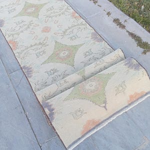 May include: A rolled-up, rectangular rug with a faded, vintage appearance. The rug features a cream-colored background with repeating geometric patterns in green, orange, and purple. The rug is placed on a gray stone surface.