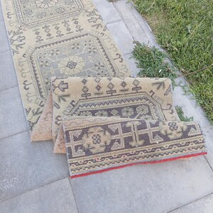 Peut inclure: Un long tapis de couloir rectangulaire présentant un motif géométrique répétitif dans des tons sourds de beige, gris et anthracite. Le tapis est replié sur lui-même, révélant un bord rouge vif le long de l'ourlet et une surface tissée.