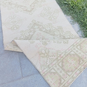Può includere: Un lungo tappeto rettangolare con base color crema e motivi geometrici a rombi ripetuti in tonalità beige tenue e rosa delicato. Il tappeto presenta una trama tessuta ed è ripiegato a un'estremità, rivelando il retro e il disegno intricato.