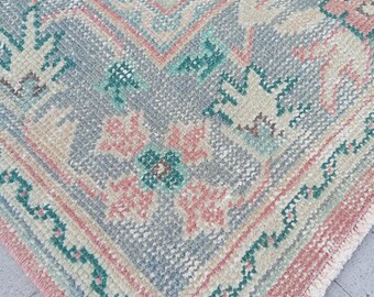 May include: Close-up of a vintage rug with a detailed geometric pattern. The rug features a color palette of soft pink, teal, beige, and gray. The design includes floral and geometric motifs, with a fringed edge.