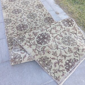 May include: A long, rectangular runner rug featuring a light beige base colour with a repeating pattern of dark brown floral motifs and intricate vine-like details. The rug is folded over at one end, revealing the woven texture and finished edges.