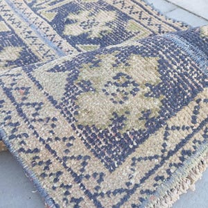 May include: A rectangular woven rug featuring a deep navy blue and muted olive green floral pattern. The rug has a thick, textured pile with a knotted construction and short, light-colored fringe along the edges, showing a folded corner.