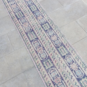 May include: A long, narrow rug with a repeating geometric pattern in shades of blue, pink, green, and cream. The rug features a central design with vertical stripes and decorative borders. The rug is placed on a light-coloured stone surface.