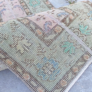 May include: A vintage rug with a distressed appearance, featuring a blend of pastel colors. The rug displays a repeating pattern of floral and geometric designs in shades of pink, green, and blue, with a brown border. The rug is partially rolled up.