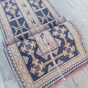 May include: A rolled-up, rectangular rug with a geometric pattern. The rug features a navy blue background with light brown, light blue, and green accents. The edges are trimmed with a red fringe. The rug is likely made of woven material.