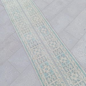 Pode incluir: Um tapete longo e estreito com um padrão geométrico repetitivo em tons de azul claro, creme e cinza. O design do tapete apresenta formas florais e geométricas estilizadas, criando uma aparência simétrica e decorativa. O tapete está colocado sobre uma superfície de ladrilhos de pedra.