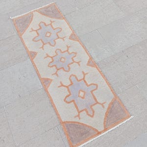 May include: A rectangular, cream-colored rug with a geometric pattern. The rug features three repeating, stylized floral designs in light blue and orange. The border and the floral elements are outlined in orange. The rug is suitable for a hallway or entryway.