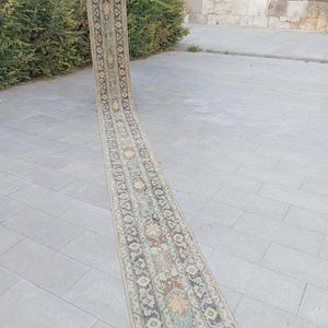 May include: A long, narrow, vintage rug with an intricate floral pattern in muted tones of beige, green, brown, and blue. The rug is displayed outdoors, showcasing its length and detailed design. The rug's edges are finished with a fringe.