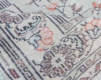 May include: A detailed close-up of a vintage rug with a faded, distressed appearance. The rug features a light green background with intricate floral and geometric patterns in shades of brown, orange, and black. The edges are slightly frayed, adding to its antique charm.