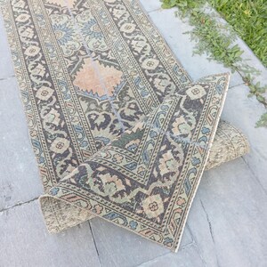 May include: A long, rectangular runner rug featuring a complex geometric and floral pattern in muted shades of charcoal gray, sage green, dusty blue, and soft peach. The rug is partially folded over, revealing its woven underside and texture.