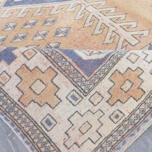 May include: A rolled-up, vintage-style rug with a geometric pattern. The rug features a cream-colored background with a repeating pattern of tan, blue, and rust-colored shapes. The edges of the rug are detailed with a blue and cream border.