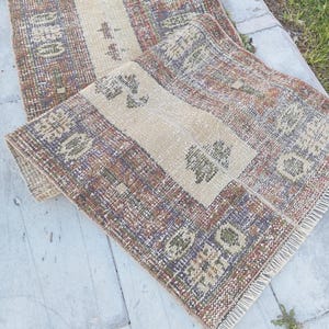 Op de afbeelding: Twee vintage tapijten met een beige middenstreep en patroonranden in bruin, paars en groen. De tapijten hebben een versleten uitstraling en een gefranjerde rand, geschikt voor interieurdecoratie.