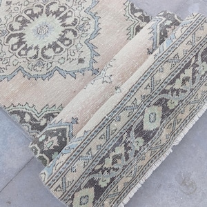 May include: A rolled-up, vintage-style rug with a beige background and intricate patterns in shades of brown, blue, and green. The rug features a central medallion design and a detailed border.