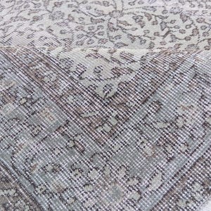 May include: Close-up of a distressed, vintage-style rug with a floral pattern. The rug features a color palette of muted greens, browns, and off-white, with a detailed, textured design. The edges of the rug are visible, showing a fringed border.