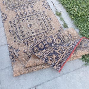 May include: A long, rectangular runner rug featuring a tan base with repeating geometric patterns in dark blue and muted brown. The rug is folded at one end, revealing a red woven edge and a dense, textured pile construction throughout the piece.