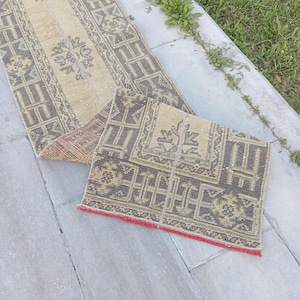 May include: A long, narrow runner rug featuring a beige base with dark grey geometric and floral patterns. The rug has a folded section revealing a red fringe edge. The design includes repeating rectangular borders and central motifs throughout.