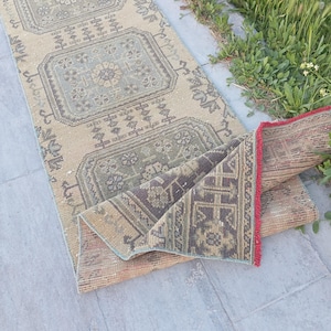 Op de afbeelding: Een beige en grijze vintage loper met geometrische patronen. Het tapijt heeft rechthoekige en bloemmotieven in tinten grijs, groen en bruin. De randen zijn afgewerkt met een rode rand. Het tapijt is gedeeltelijk opgerold en toont de textuur en het ontwerp.