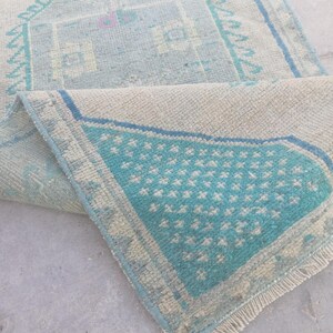 May include: A woven rug featuring a light beige base with geometric patterns in shades of turquoise and blue. The rug has a folded corner revealing a textured underside and a short, light-colored fringe along the edge of the rectangular textile.