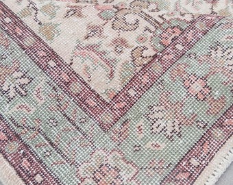 May include: A vintage-style rug with a detailed floral pattern. The rug features a cream-colored background with accents of pink, green, and burgundy. The border has a geometric design in shades of green and burgundy. The rug has a fringed edge.