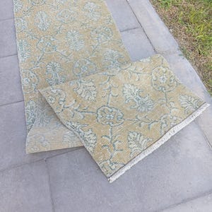 May include: A long, rectangular runner rug featuring a mustard yellow base colour with a repeating floral and leaf pattern in a muted teal blue. The rug has a woven texture and short white fringe along the edges, showing a folded section.