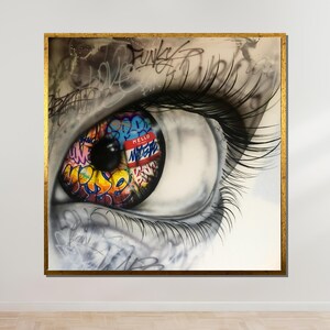 Puede incluir: Una obra de arte enmarcada que representa un ojo detallado con largas pestañas. El iris es un collage vibrante de arte graffiti, incluyendo la palabra "HELLO" en una etiqueta roja. La obra está sobre un fondo blanco con un marco dorado.
