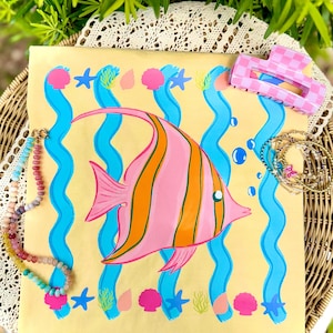 Può includere: Una borsa tote gialla con un'illustrazione di pesce rosa e arancione, linee ondulate blu e accenti a tema marino. Sono visibili anche un fermaglio per capelli rosa e bianco a scacchi e gioielli di perline colorate.