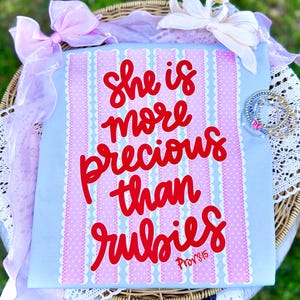 Puede incluir: Un letrero decorativo azul claro con el texto "she is more precious than rubies" en escritura roja. El letrero tiene detalles de rayas rosas y blancas y está decorado con lazos y pulseras.