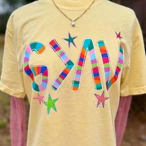 May include: Yellow t-shirt with the word "GAV" in colorful, patchwork-style letters. The shirt features several star-shaped designs in various colors. A gold necklace with a pendant is visible.