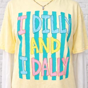 May include: Yellow short-sleeved t-shirt with the text "I DILLY AND I DALLY" in a colorful, striped design. The shirt is layered over a white long-sleeved shirt. A gold necklace with a small pendant is visible.