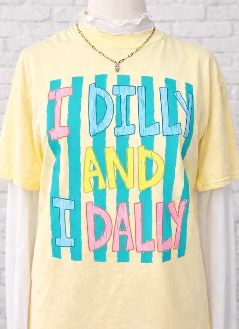 K&ouml;nnte beinhalten: Hellgelbes T-Shirt mit dem Text "I DILLY AND I DALLY" in einem farbenfrohen, gestreiften Design. Das T-Shirt ist &uuml;ber einem wei&szlig;en Langarmshirt geschichtet. Eine goldene Halskette mit einem kleinen Anh&auml;nger ist sichtbar.