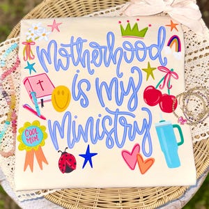 Può includere: T-shirt color crema con la scritta blu "Motherhood is my Ministry". La maglietta è decorata con illustrazioni colorate tra cui una corona, ciliegie, un arcobaleno e una faccina sorridente. È presente anche un nastro "Cool Mom".